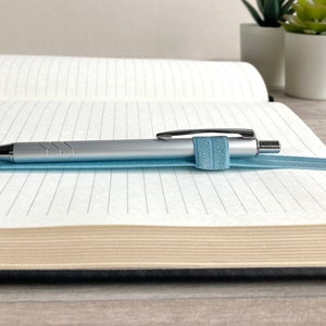 May include: A silver pen with a blue elastic band is resting on a lined notebook with a cream-colored cover.