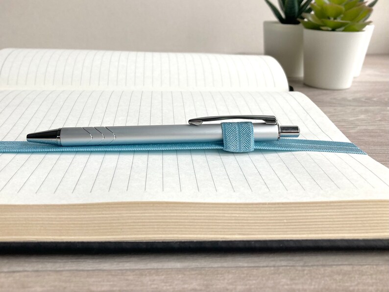 May include: A silver pen with a blue elastic band is resting on a lined notebook with a cream-colored cover.