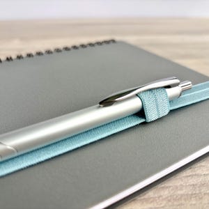 Puede incluir: Un bol&iacute;grafo plateado con una banda el&aacute;stica azul claro unida a una libreta de espiral gris.