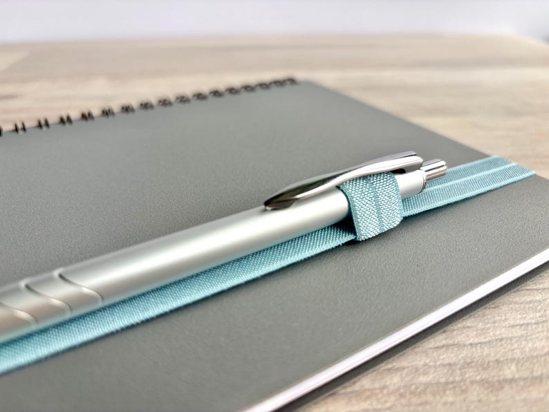 Puede incluir: Un bol&iacute;grafo plateado con una banda el&aacute;stica azul claro unida a una libreta de espiral gris.