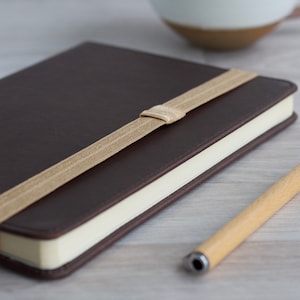 May include: A brown leather-bound journal with a tan elastic closure and a wooden pen.