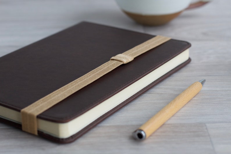 May include: A brown leather-bound journal with a tan elastic closure and a wooden pen.