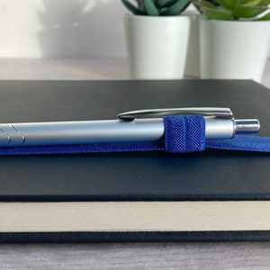May include: A silver pen with a blue elastic band holding it to a black notebook.