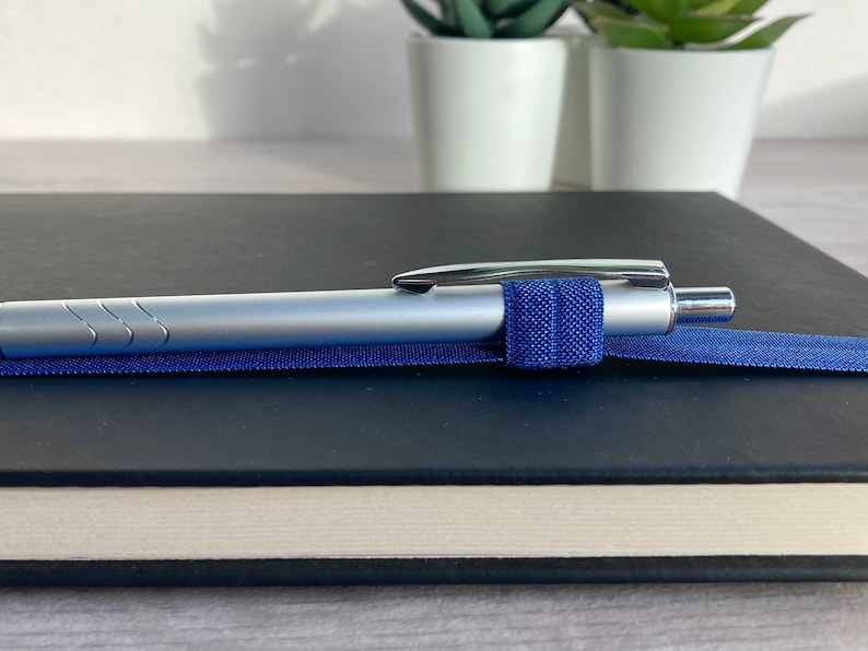 May include: A silver pen with a blue elastic band holding it to a black notebook.