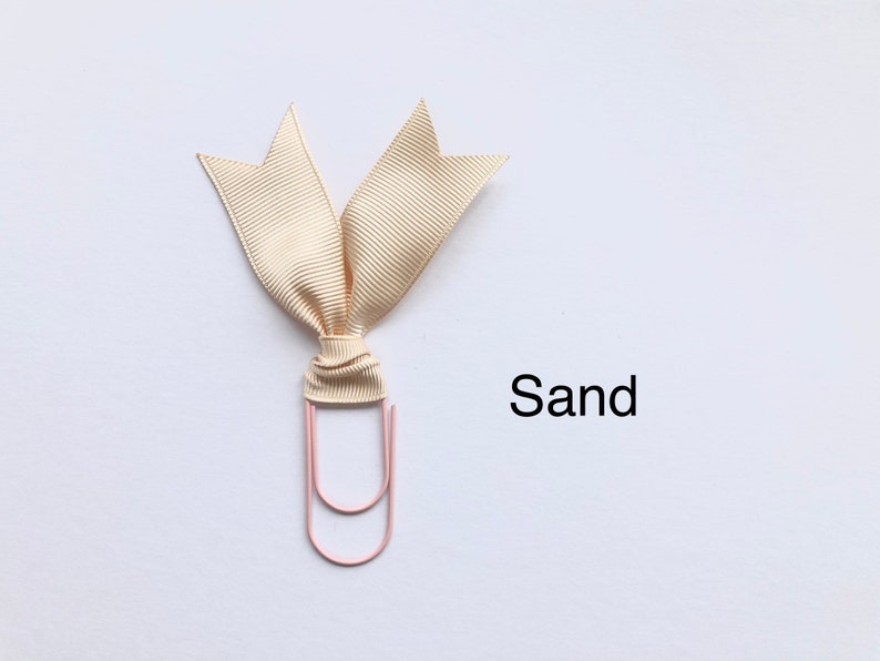 Może przedstawiać: Beżowa wstążka zawiązana w kokardę wok&oacute;ł r&oacute;żowego spinacza. Tekst "Sand" jest napisany pod wstążką.