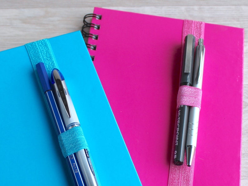 May include: Two notebooks with elastic bands holding pens. The notebook on the left is blue with two pens, one blue and one silver. The notebook on the right is pink with two pens, one black and one silver. The black pen has the text "Hi-Tecpoint 0.8" printed on it.