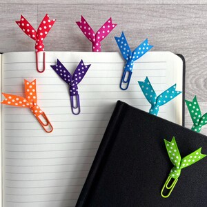 May include: An open notebook with several colorful polka-dot ribbon bookmarks attached to paper clips. The bookmarks are red, pink, blue, green, orange, and purple. A black notebook is also visible.