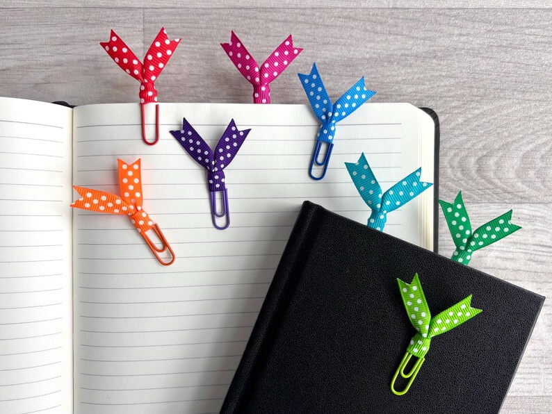 May include: An open notebook with several colorful polka-dot ribbon bookmarks attached to paper clips. The bookmarks are red, pink, blue, green, orange, and purple. A black notebook is also visible.
