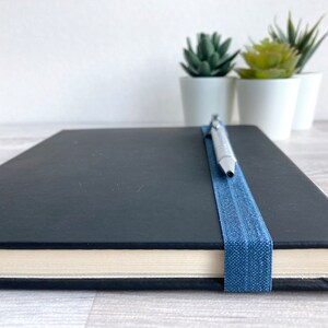 May include: A black notebook with a blue elastic band and a silver pen resting on top. The notebook is closed and sitting on a light wood surface.
