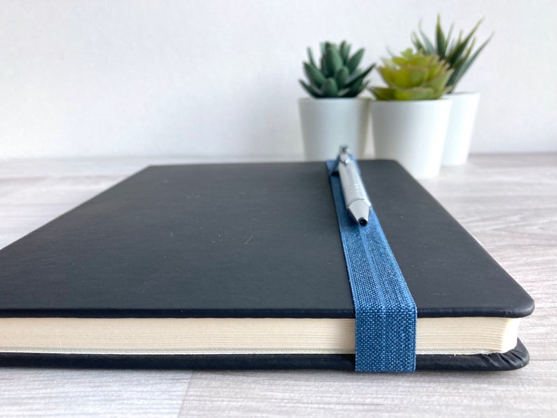 May include: A black notebook with a blue elastic band and a silver pen resting on top. The notebook is closed and sitting on a light wood surface.