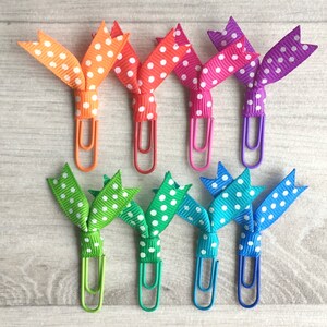 May include: A collection of colorful paper clips with polka dot ribbon bows. The paper clips are in various colors, including orange, red, pink, purple, green, teal, and blue. The ribbon is white with polka dots.