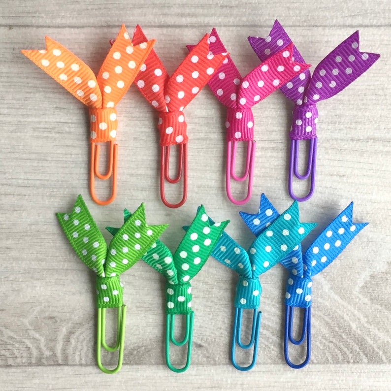 May include: A collection of colorful paper clips with polka dot ribbon bows. The paper clips are in various colors, including orange, red, pink, purple, green, teal, and blue. The ribbon is white with polka dots.