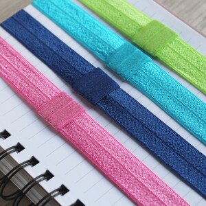 May include: Five colorful elastic hair ties in pink, blue, green, and navy blue. The hair ties are arranged on a white notebook with black spiral binding.