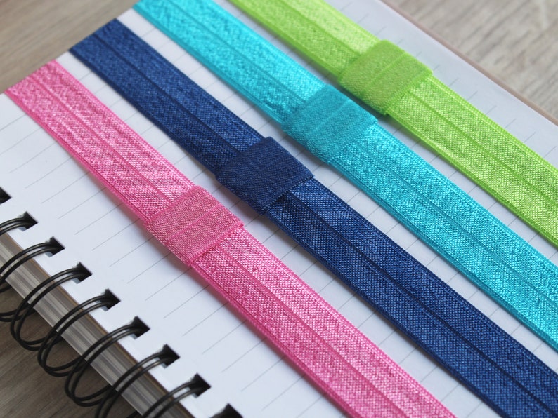 May include: Five colorful elastic hair ties in pink, blue, green, and navy blue. The hair ties are arranged on a white notebook with black spiral binding.