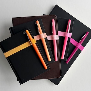 May include: A collection of black and brown notebooks with colorful pens. The notebooks are secured with elastic bands in shades of orange, pink, and magenta. The pens are in matching colors, creating a coordinated stationery set.