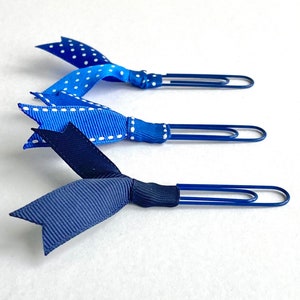 May include: Three blue paper clips with blue and white ribbon bows. The ribbons are tied in a bow and the ends are tucked into the paper clip.