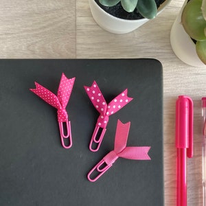 May include: Three pink paper clips with decorative ribbon accents. Two clips have white polka dots and one has a solid pink ribbon. The clips are on a black surface.