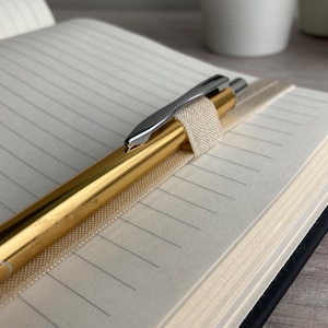 May include: A gold pen with a silver clip is placed in a notebook with lined pages. The notebook is closed and has a black cover.