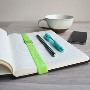 May include: An open notebook with lined pages, a green elastic band bookmark, a blue pen, and a black pencil on a wooden table. A white ceramic mug and a black smartphone are also on the table.