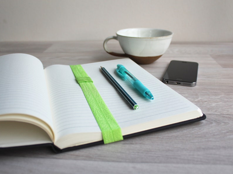 May include: An open notebook with lined pages, a green elastic band bookmark, a blue pen, and a black pencil on a wooden table. A white ceramic mug and a black smartphone are also on the table.
