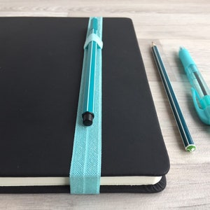 May include: A black notebook with a teal pen secured by a light blue elastic band. A teal pen and a pencil are lying on a light wood surface.