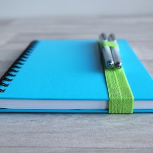 May include: A blue spiral bound notebook with a green elastic band holding two silver pens in place.