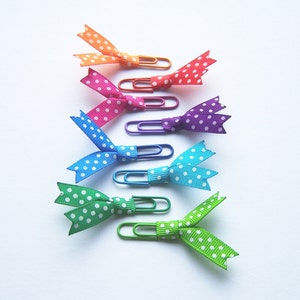 May include: A collection of colorful paper clips, each adorned with a polka-dot ribbon bow. The bows are in shades of orange, red, pink, blue, teal, green, and lime green. The paper clips are in matching colors.