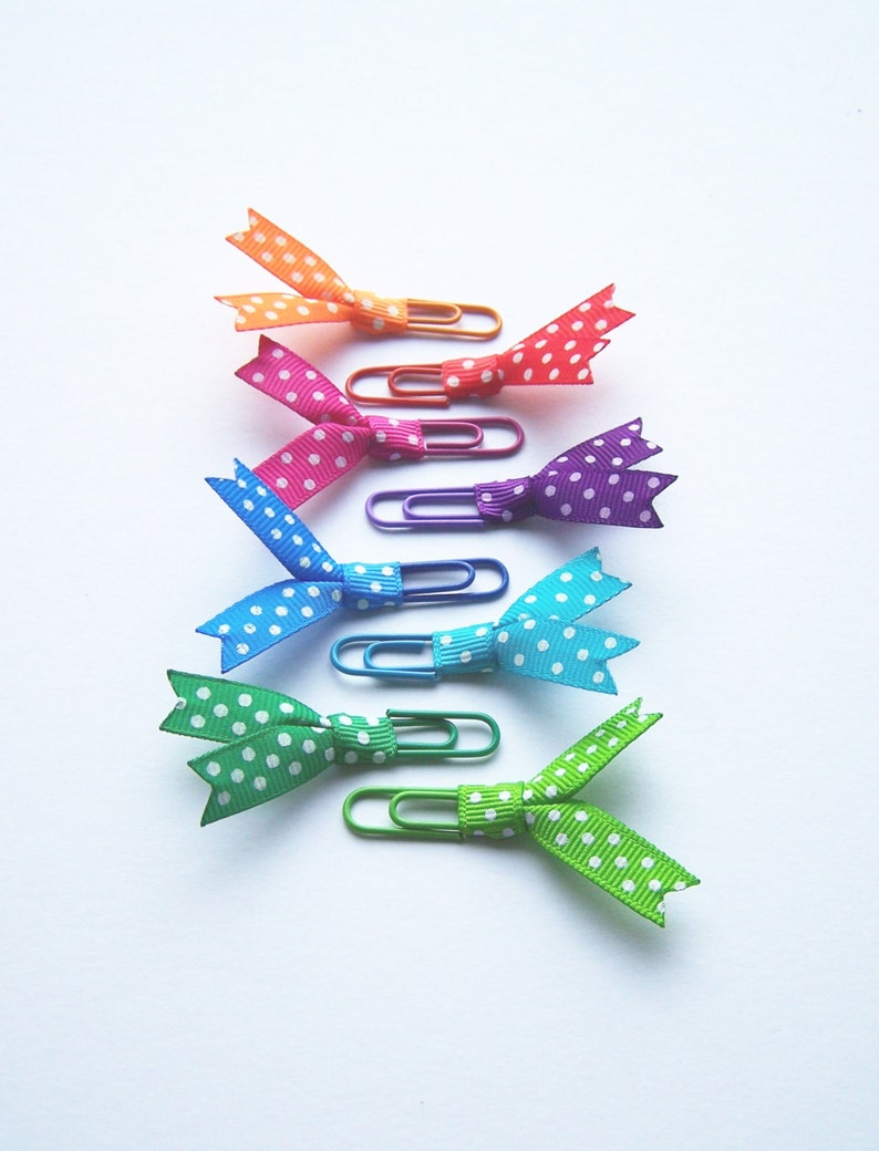 May include: A collection of colorful paper clips, each adorned with a polka-dot ribbon bow. The bows are in shades of orange, red, pink, blue, teal, green, and lime green. The paper clips are in matching colors.
