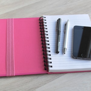 May include: A pink notebook with a black spiral binding and a black elastic band closure. The notebook is open to reveal lined white pages. A black pen, a silver pen, and a black smartphone are on the open pages.