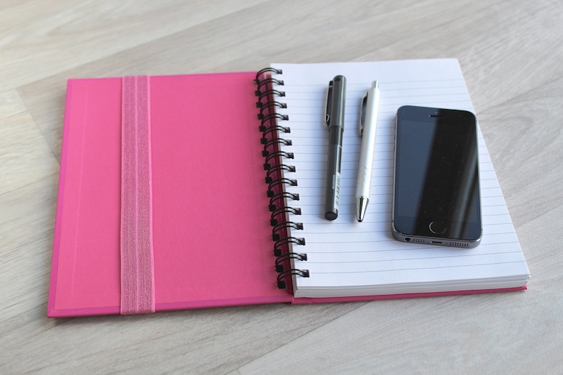 May include: A pink notebook with a black spiral binding and a black elastic band closure. The notebook is open to reveal lined white pages. A black pen, a silver pen, and a black smartphone are on the open pages.
