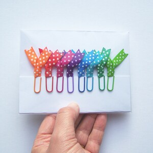 May include: A set of colorful paper clips with polka dot ribbon bows in a rainbow of colors: orange, red, pink, purple, blue, and green. The paper clips are held in a white card.