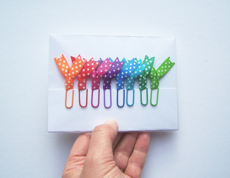 May include: A set of colorful paper clips with polka dot ribbon bows in a rainbow of colors: orange, red, pink, purple, blue, and green. The paper clips are held in a white card.