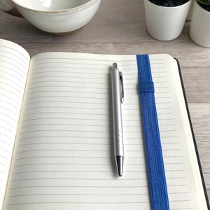 May include: An open lined notebook with a blue elastic band bookmark and a silver pen lying on the open pages.