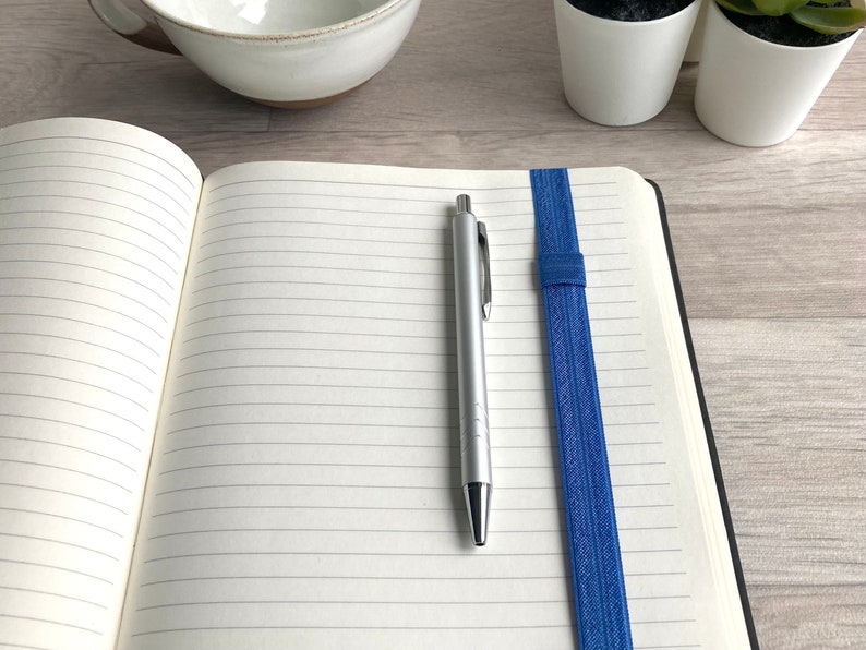 May include: An open lined notebook with a blue elastic band bookmark and a silver pen lying on the open pages.