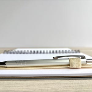 Puede incluir: Un bol&iacute;grafo plateado con una banda el&aacute;stica beige descansa sobre un cuaderno blanco con encuadernaci&oacute;n en espiral.