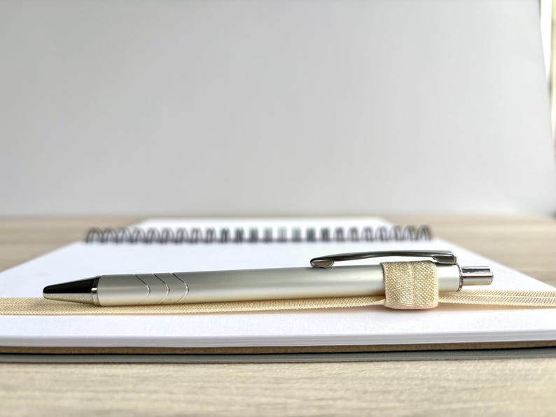 Puede incluir: Un bol&iacute;grafo plateado con una banda el&aacute;stica beige descansa sobre un cuaderno blanco con encuadernaci&oacute;n en espiral.