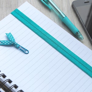 May include: A white lined notebook with a turquoise elastic band and a turquoise ribbon bookmark with white polka dots. A turquoise pen and a silver paperclip are on the notebook.