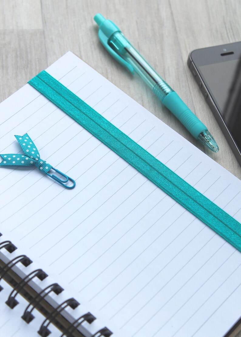 May include: A white lined notebook with a turquoise elastic band and a turquoise ribbon bookmark with white polka dots. A turquoise pen and a silver paperclip are on the notebook.