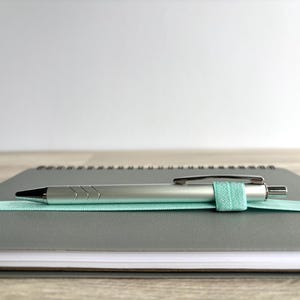 May include: A grey spiral bound notebook with a silver pen and a light blue elastic band holding the pen in place.