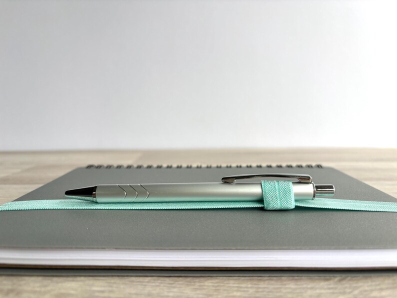 May include: A grey spiral bound notebook with a silver pen and a light blue elastic band holding the pen in place.