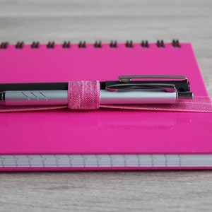 May include: A pink spiral bound notebook with two pens secured with a pink ribbon. The pens are silver and black.