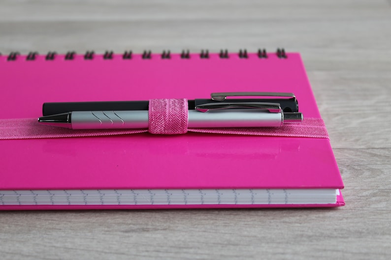 May include: A pink spiral bound notebook with two pens secured with a pink ribbon. The pens are silver and black.
