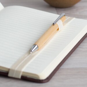 May include: A brown leather bound journal with a white ribbon bookmark and a wooden pen with a silver tip.