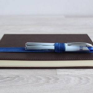May include: A brown leather-bound journal with a blue ribbon bookmark and two silver pens secured with a blue elastic band.