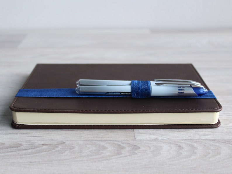 May include: A brown leather-bound journal with a blue ribbon bookmark and two silver pens secured with a blue elastic band.