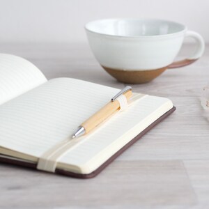 May include: A blank notebook with a brown cover, a wooden pen with a silver clip, and a white ceramic mug on a light wood surface.