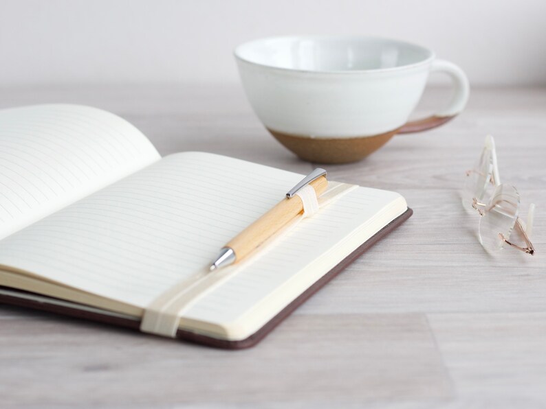 May include: A blank notebook with a brown cover, a wooden pen with a silver clip, and a white ceramic mug on a light wood surface.