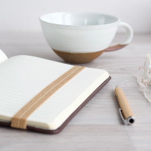 May include: A brown leather bound journal with a beige fabric bookmark. The journal is open to a blank page with lined paper. A white ceramic mug and a pair of clear glasses are also in the image.