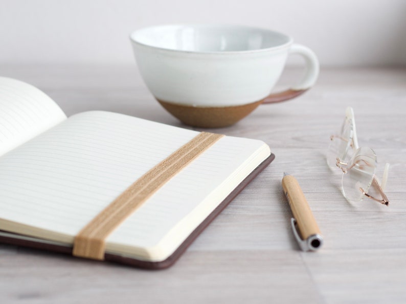 May include: A brown leather bound journal with a beige fabric bookmark. The journal is open to a blank page with lined paper. A white ceramic mug and a pair of clear glasses are also in the image.