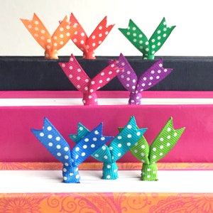 May include: A collection of colorful ribbon bookmarks with white polka dots. The bookmarks are in various colors, including orange, red, green, pink, purple, blue, and teal. They are placed on top of books with different colored spines.